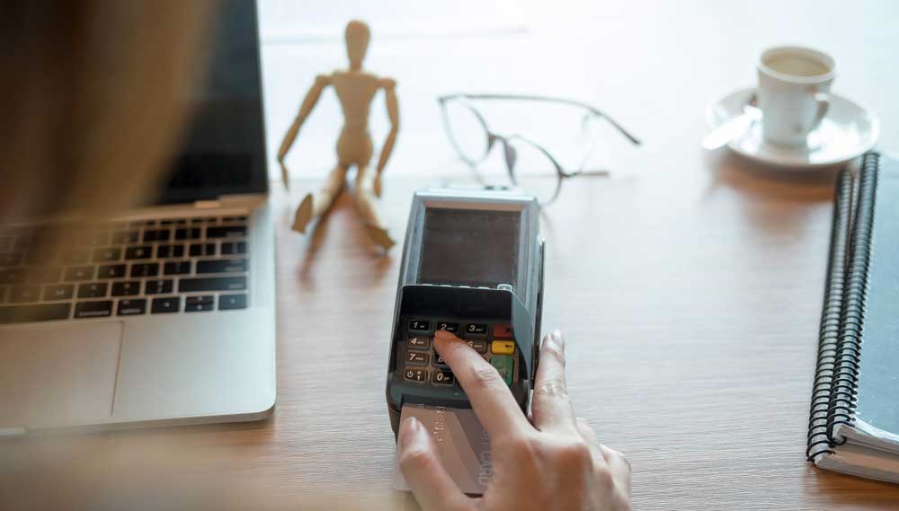 A person's hand using a card reader to process a payment with a small wooden mannequin, eyeglasses, a notepad, and a cup of coffee in the background, signifying a busy work environment or a point-of-sale setting.